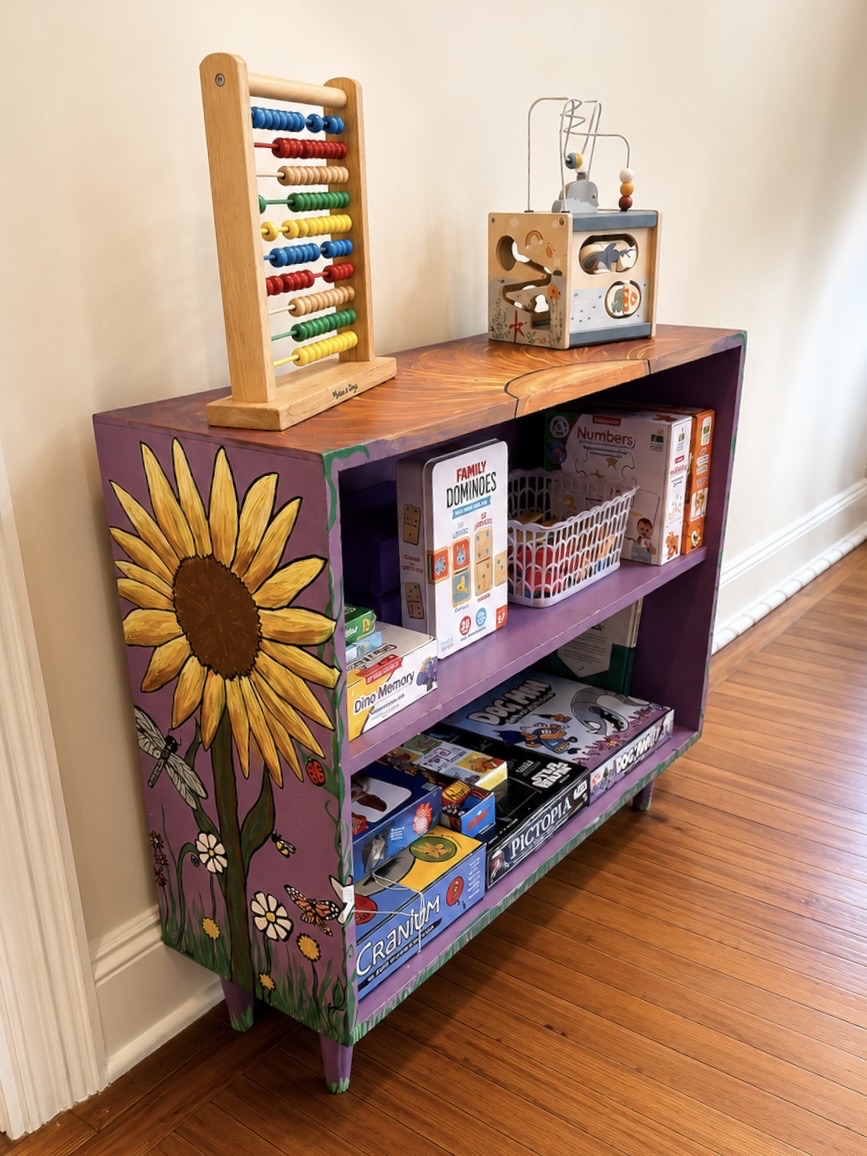 A hand painted purple bookshelf with a sunflower on the side, holding board games, a wooden abacus and a wooden activity cube.