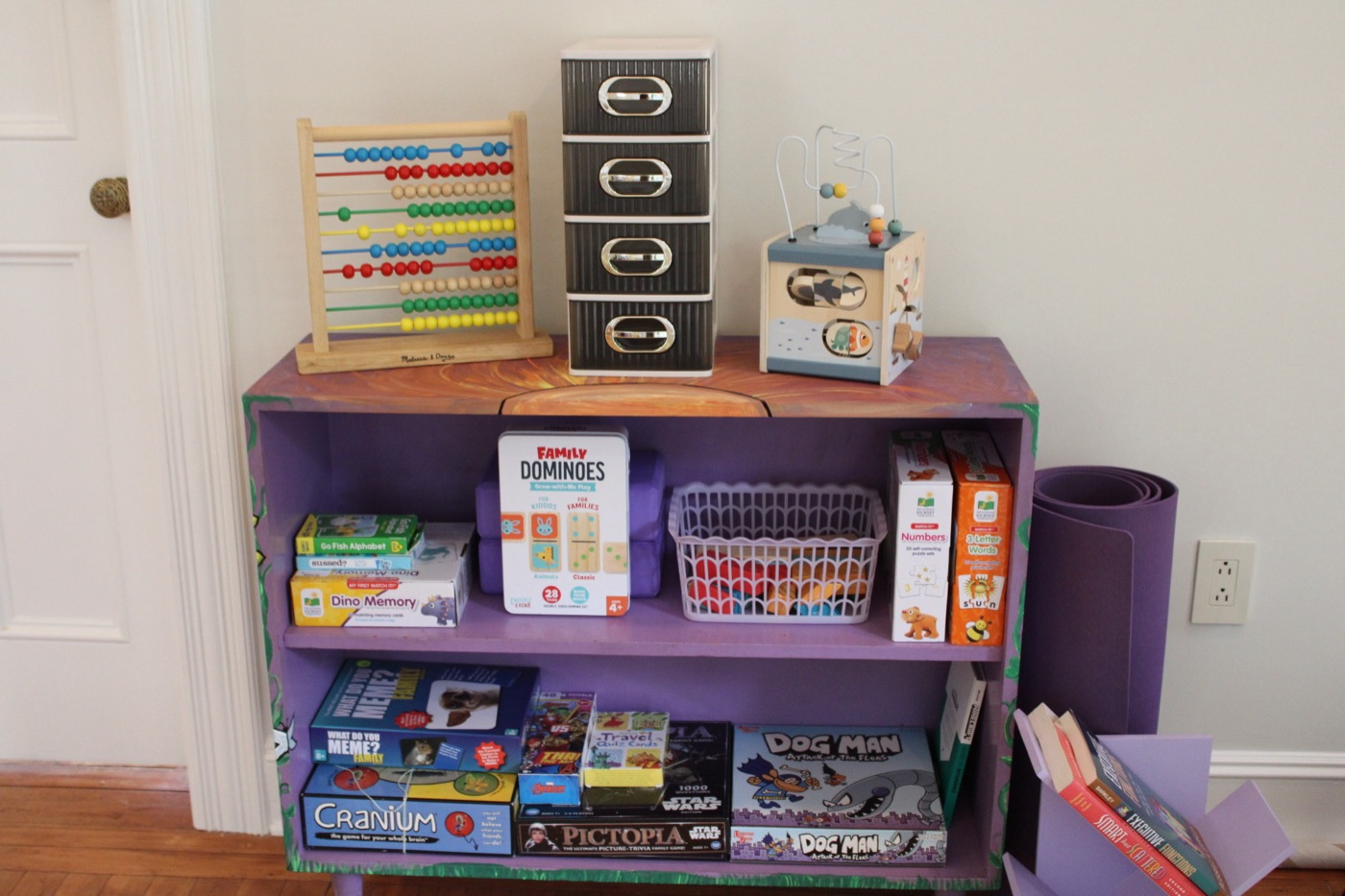 A hand painted purple bookshelf holding board games, dominoes, memory games, a wooden abacus and a basket of counting animals.