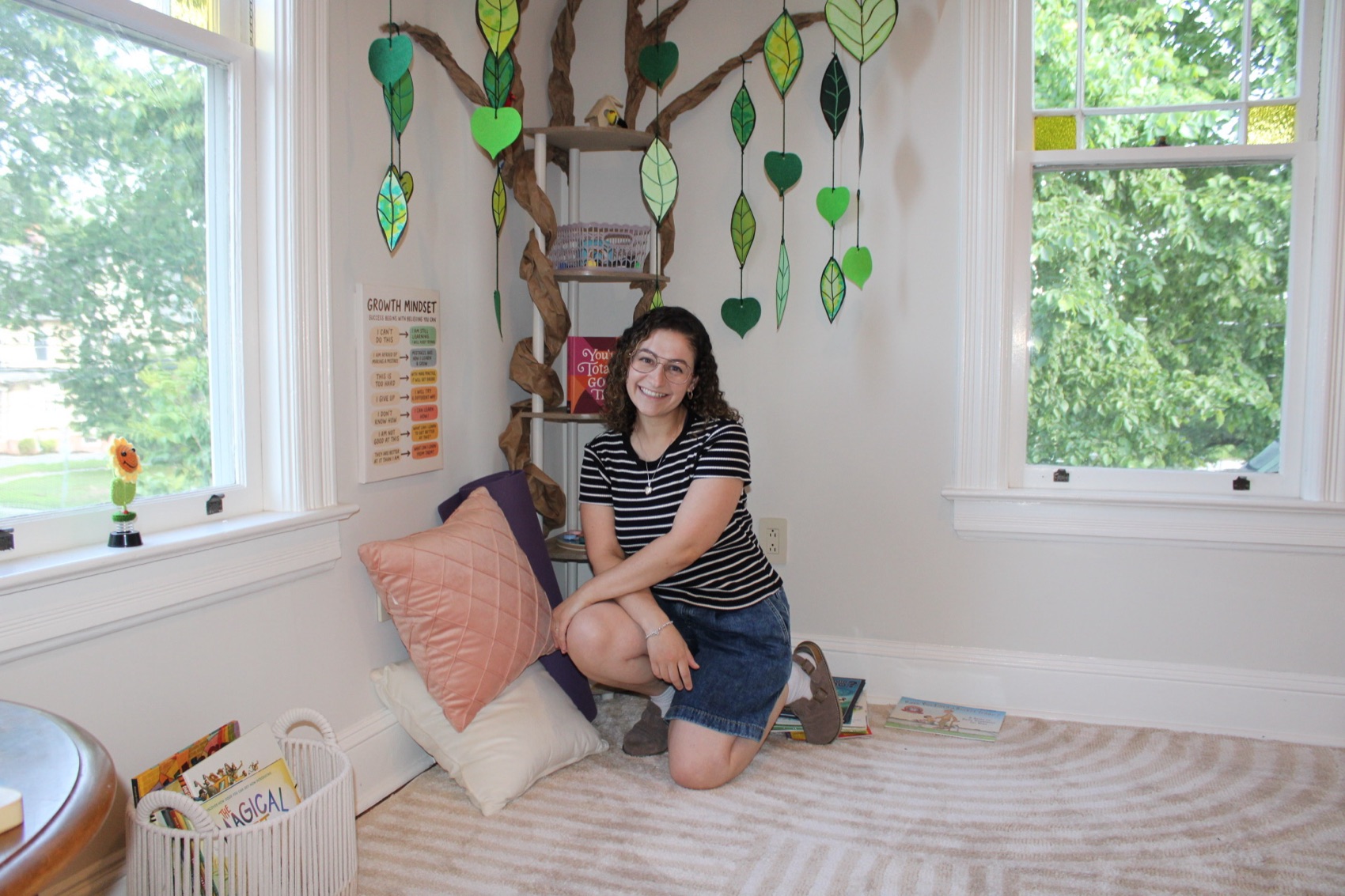 Linda kneeling in the reading nook of the Grow Space room, smiling, with the paper tree and hanging leaves behind her.