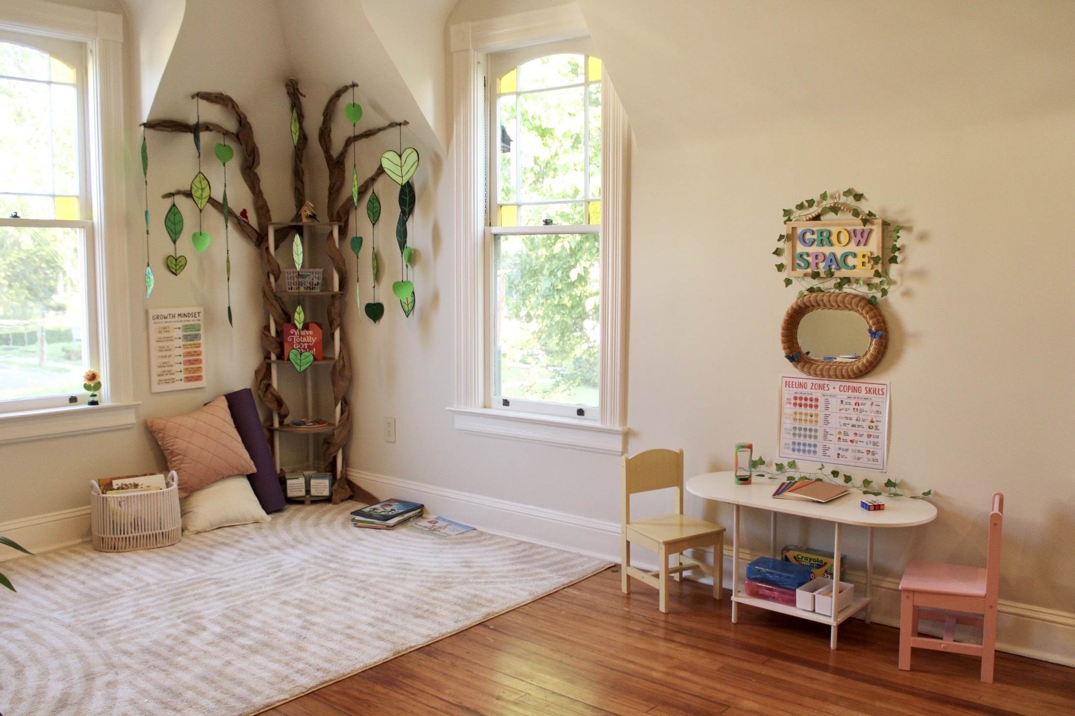 The Grow Space room. A braided paper tree climbs the corner with green paper leaves and hearts hanging from its branches, beside a cushioned reading nook and a small work table under a feelings chart.